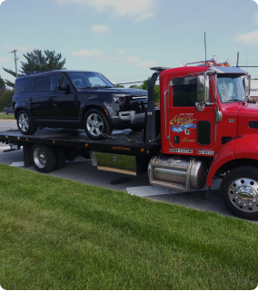 A red Azar light-duty flatbed tow truck with a black SUV chained to its bed.