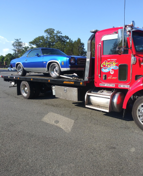 An Azar light-duty flatbed tow truck with a classic blue car chained to its bed.