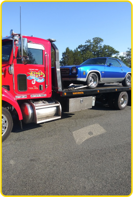 An Azar light-duty flatbed tow truck with a classic blue car chained to its bed.