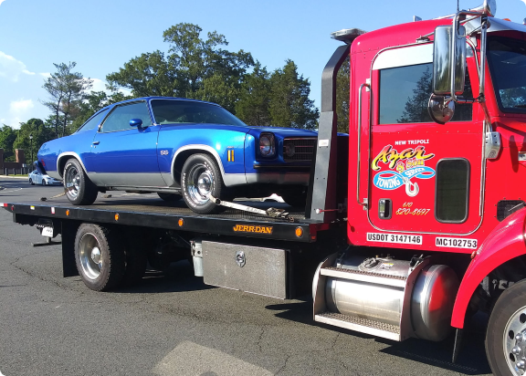 An Azar light-duty flatbed tow truck with a classic blue car chained to its bed.