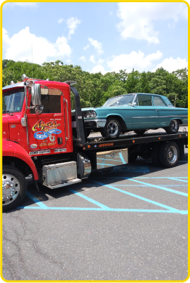 A classic teal muscle car on the flatbed of a red Azar tow truck in a parking lot.