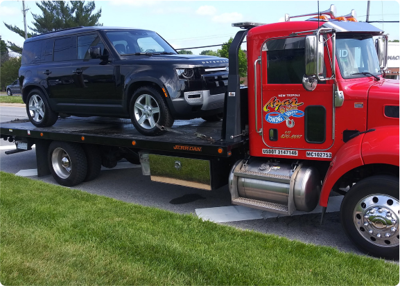 A red Azar light-duty flatbed tow truck with a black SUV chained to its bed