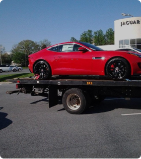 A red classic luxury car parked on the back of a flatbed transport truck in front of a Jaguar dealership.