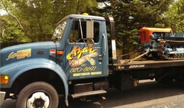 A blue-cabbed Azar tow truck hauls an orange and blue treaded tractor