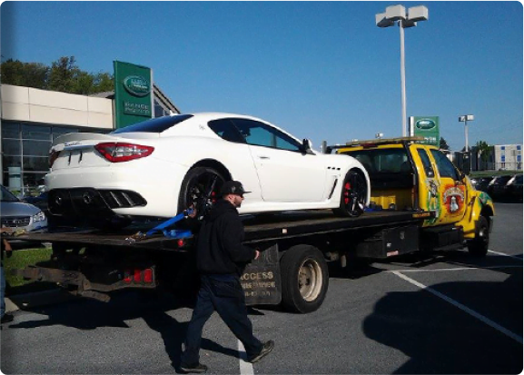 Rear view of a yellow flatbed tow truck hauling a white luxury car at a Rangerover dealership