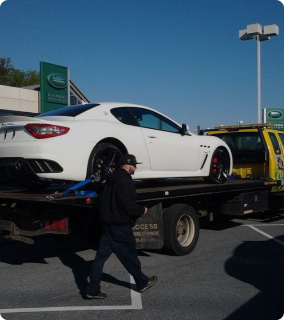 Rear view of a yellow flatbed tow truck hauling a white Maserati at a Rangerover dealership