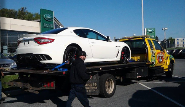 Rear view of a yellow flatbed tow truck hauling a white luxury car at a Rangerover dealership