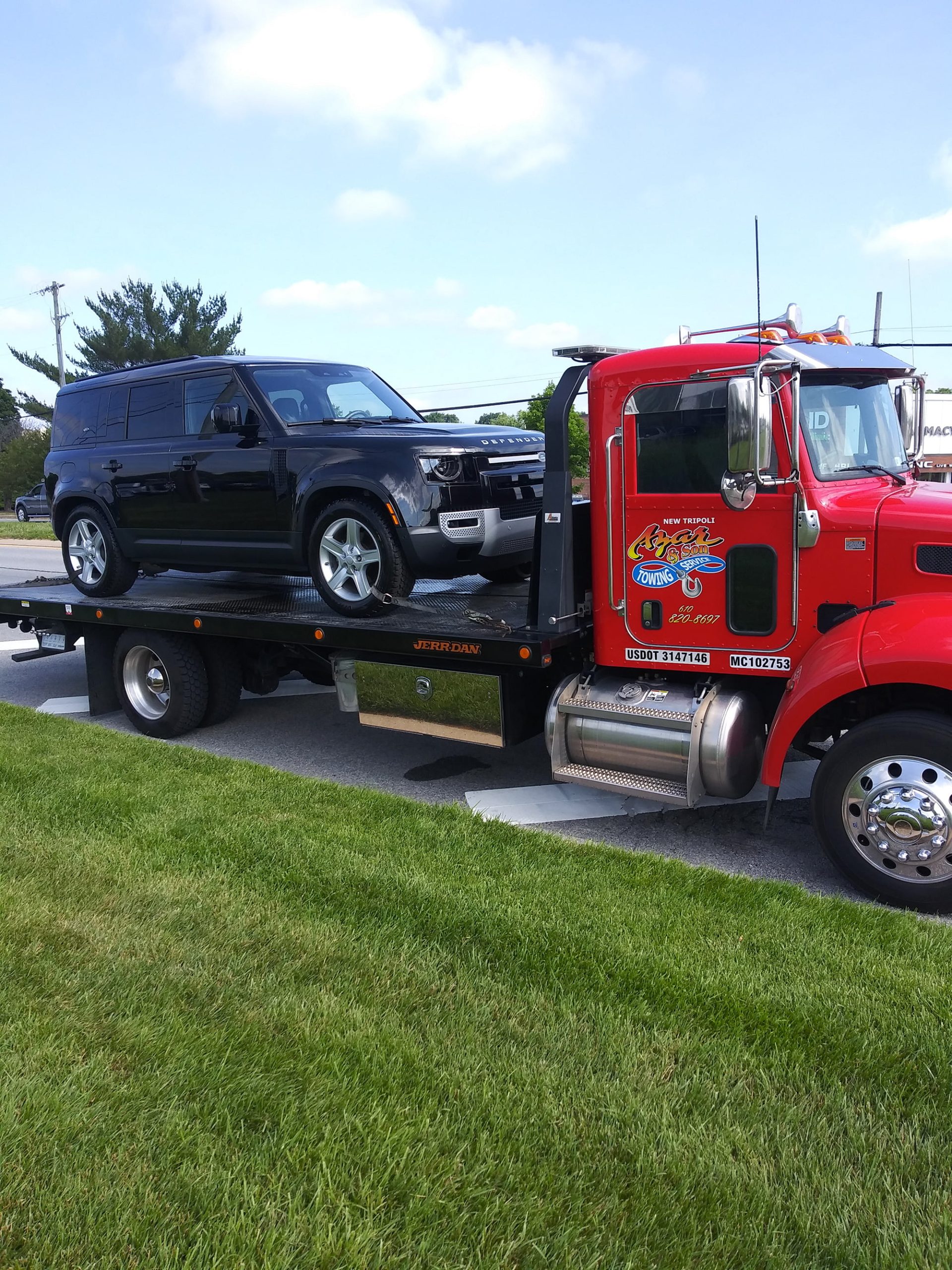 A red Azar light-duty flatbed tow truck with a black SUV chained to its bed