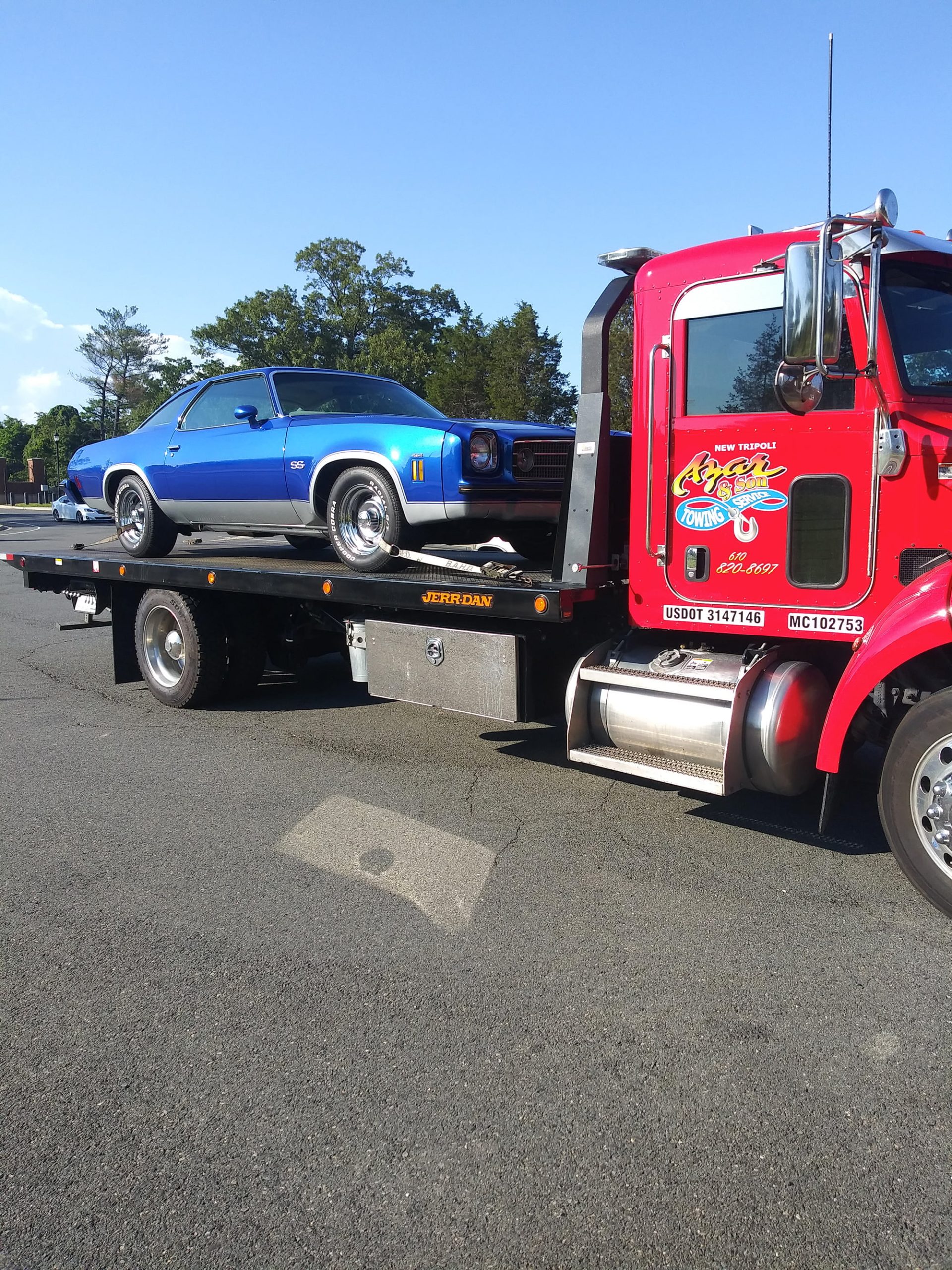 An Azar light-duty flatbed tow truck with a classic blue car chained to its bed.