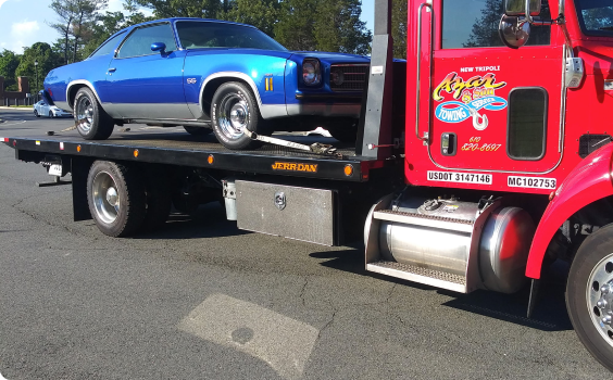 An Azar light-duty flatbed tow truck with a classic blue car chained to its bed.