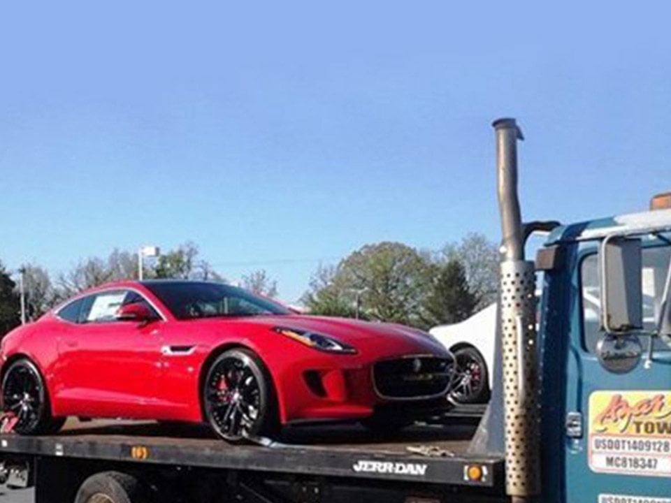 A luxury red car parked on the back of a blue-cabbed flatbed transport truck on a car lot.