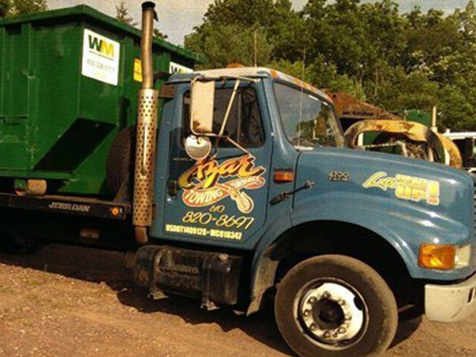 a blue-cabbed Azar tow truck hauls a green Waste Management dumpster